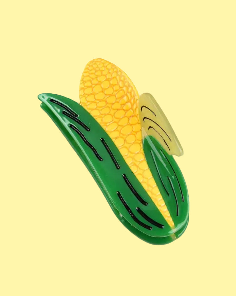 A novelty hair claw clip shaped like a corn cob, featuring a yellow textured cob and green leaves, displayed on a white surface with a brown wooden tray in the background.
